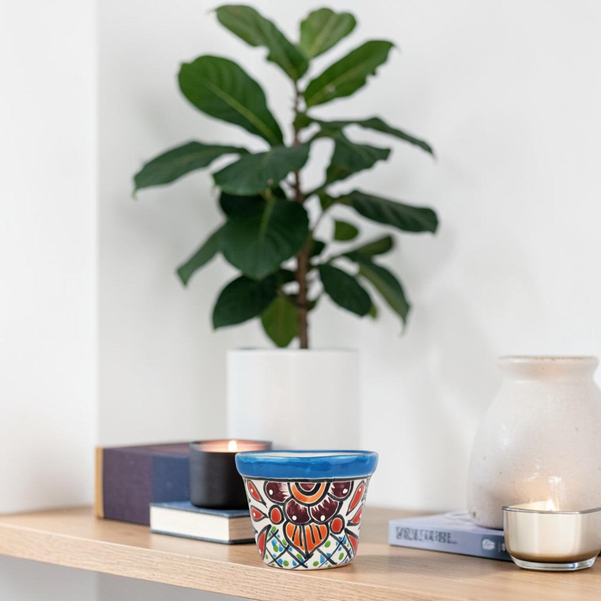 A small hand-painted ceramic pot with a colorful mandala pattern and a blue rim, displayed on a wooden shelf next to candles and a potted fiddle-leaf fig plant.