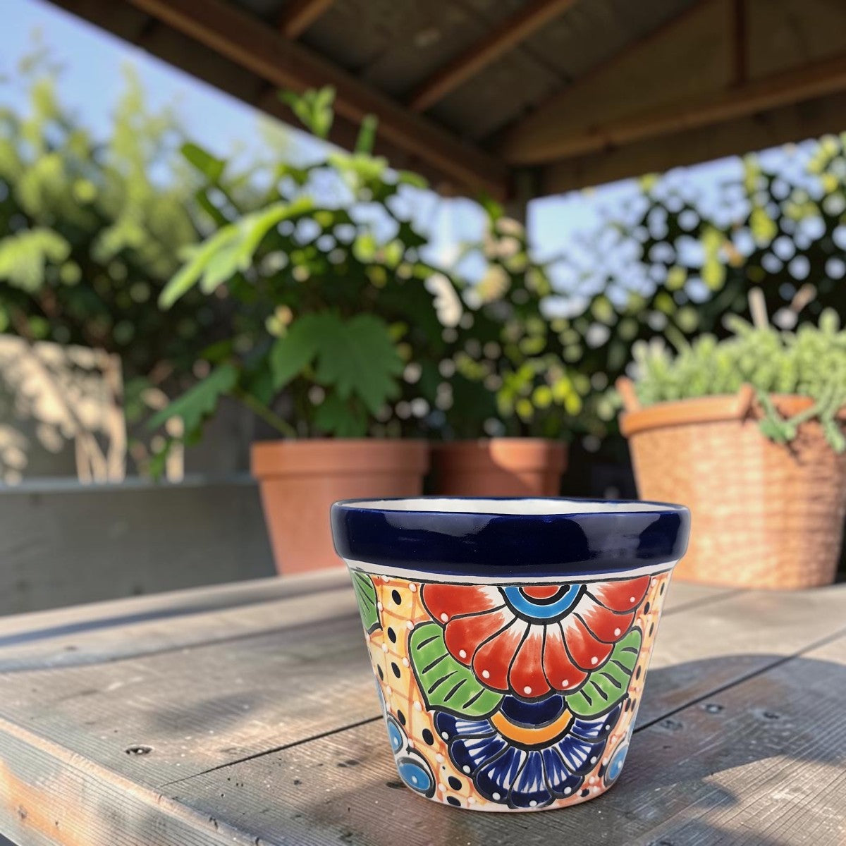 A colorful hand-painted ceramic planter pot with a navy blue rim sits on a weathered wooden table in an outdoor garden setting.