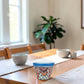 A colorful hand-painted ceramic planter with a blue rim sits on a wooden dining table in a bright, modern room with a blurred plant and chairs in the background.