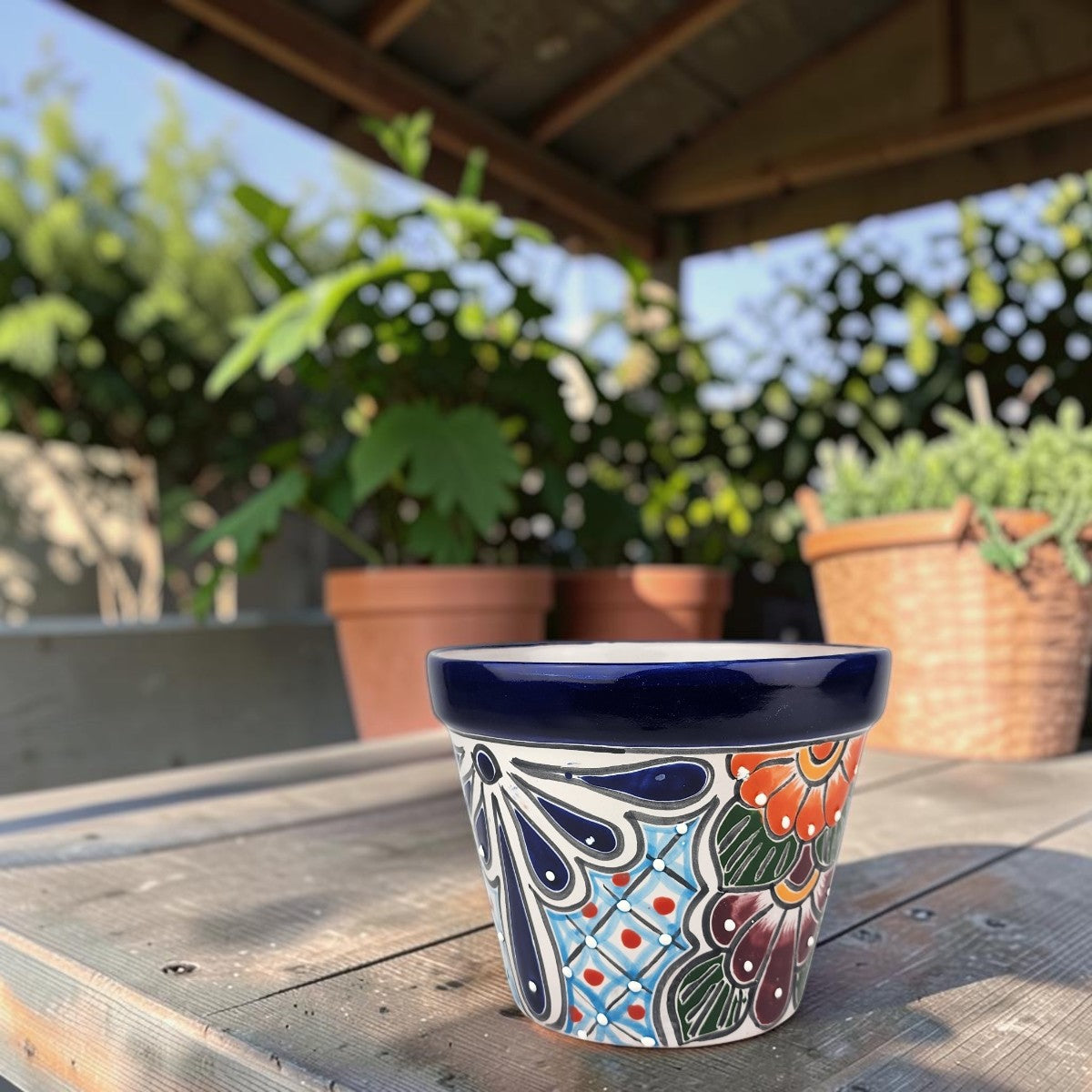 A small hand-painted ceramic planter pot with a Mexican folk art design, sitting on a weathered wooden table in an outdoor garden setting.