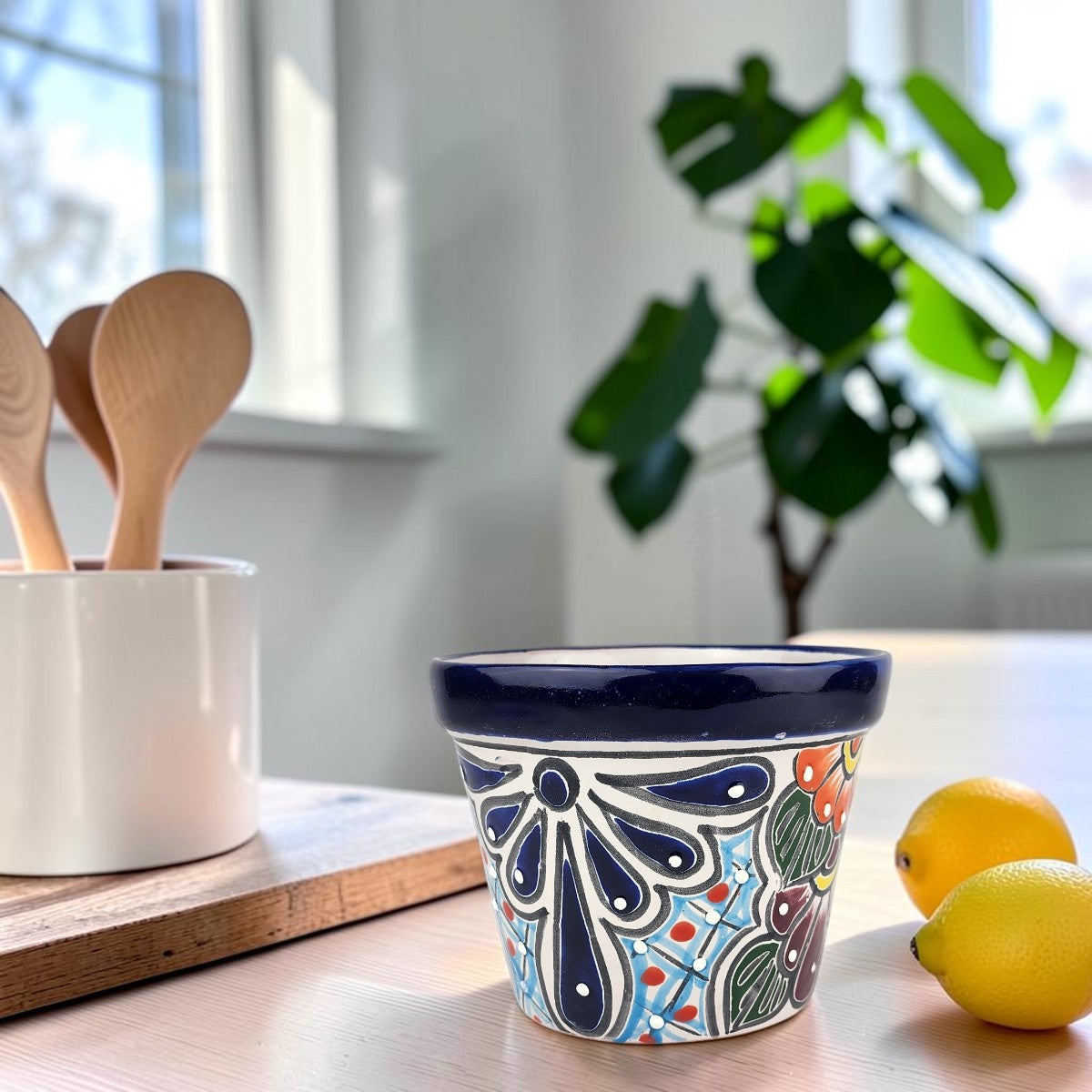 A small hand-painted ceramic pot with a navy blue rim and a colorful floral pattern, styled on a wooden table next to lemons and a white ceramic utensil holder in a bright kitchen setting.
