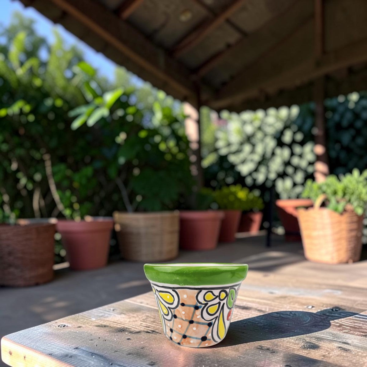 A small hand-painted ceramic planter pot with a green rim and a colorful floral pattern, sitting on a wooden table in a sunny garden patio setting.