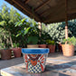 A hand-painted ceramic planter pot with a vibrant blue rim and a colorful Mexican-style floral pattern, sitting on a wooden table in a sunny garden patio setting.