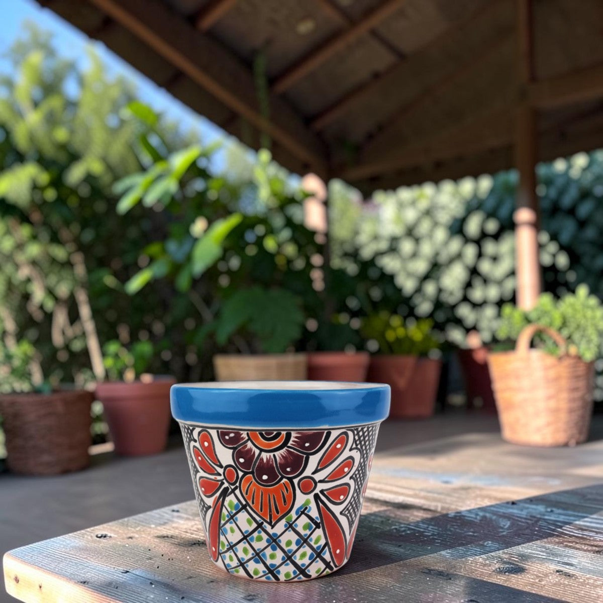 A hand-painted ceramic planter pot with a vibrant blue rim and a colorful Mexican-style floral pattern, sitting on a wooden table in a sunny garden patio setting.