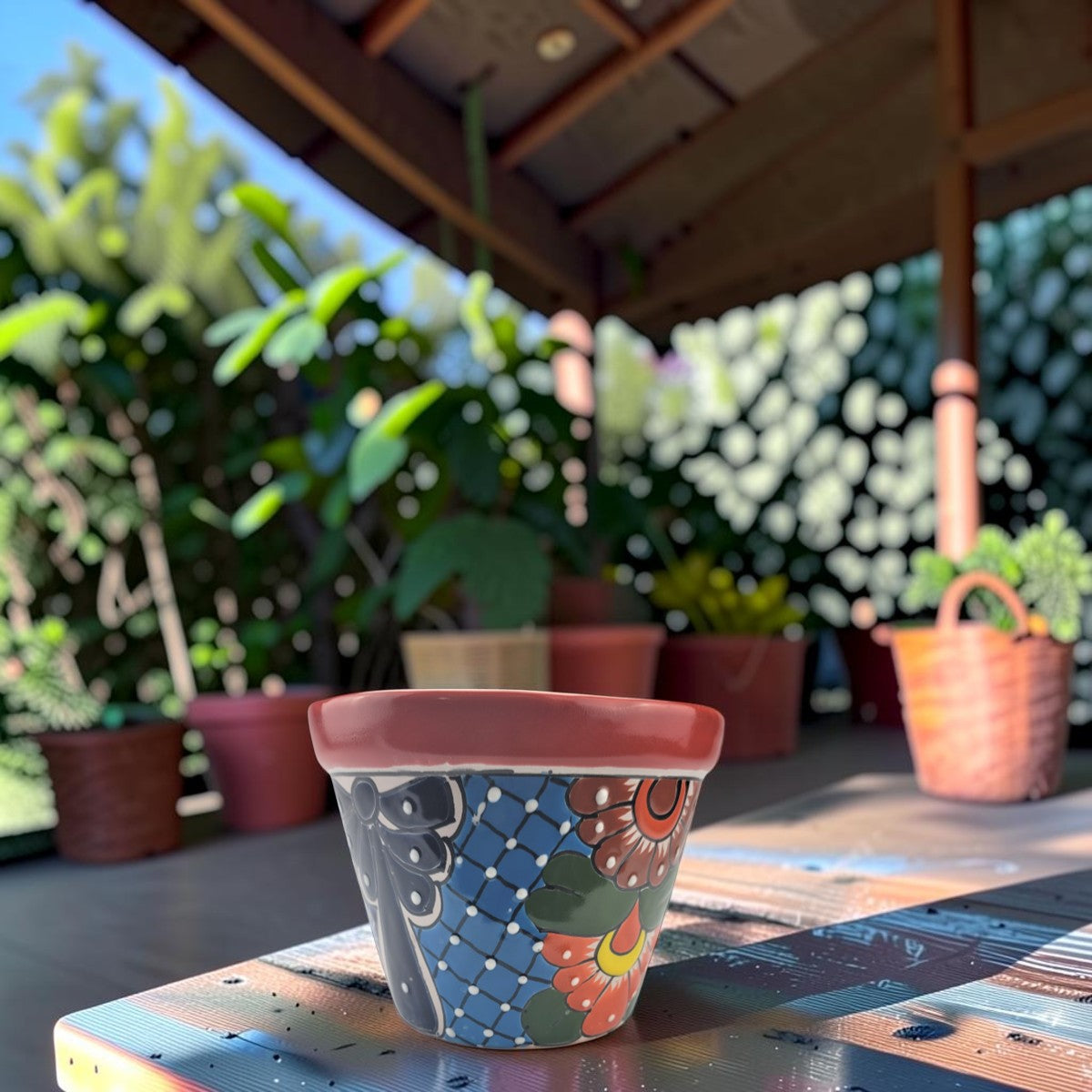 A small hand-painted ceramic planter pot with a red-colored rim and a colorful floral and geometric pattern, sitting on a wooden table under a covered patio with potted plants in the background.