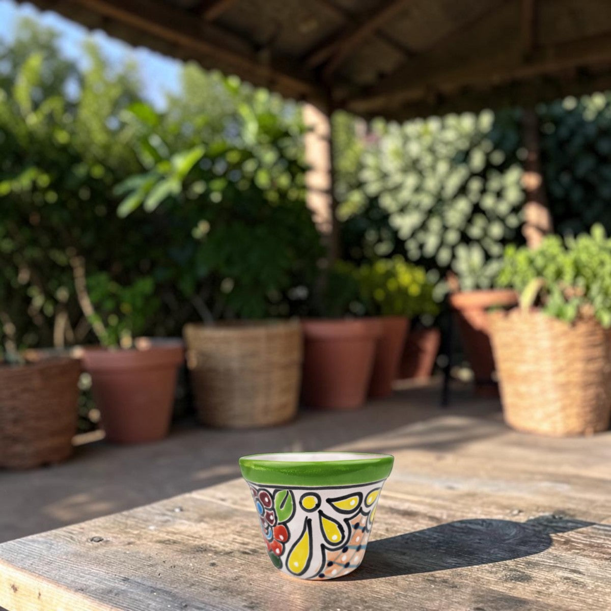 A small hand-painted ceramic planter with a green rim and colorful floral patterns sits on a rustic wooden table in an outdoor patio setting with potted plants in the background.