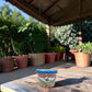 A small hand-painted ceramic planter pot with a blue rim and orange, green, and white floral scrollwork, sitting on a wooden table under a covered patio with potted plants in the background.