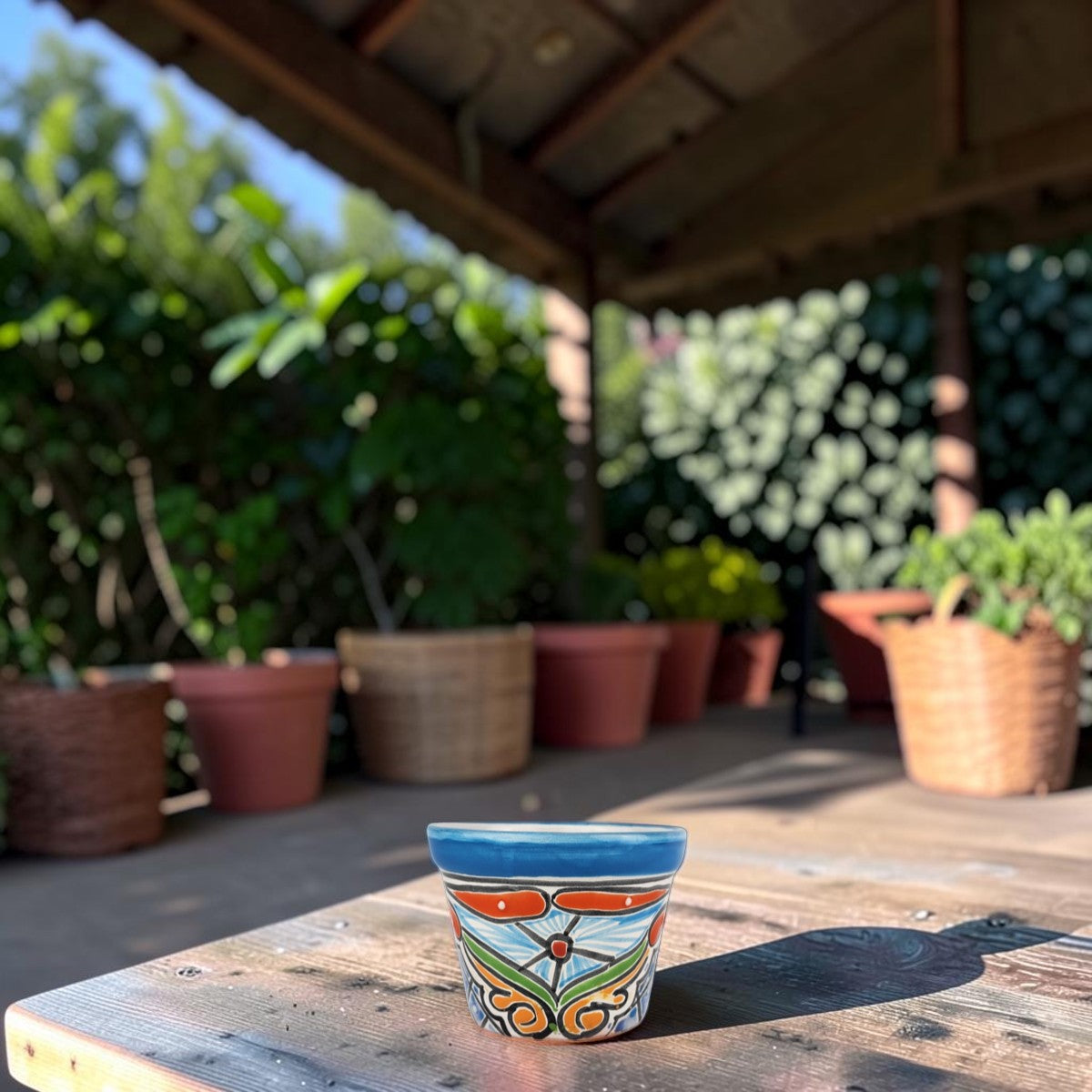 A small hand-painted ceramic planter pot with a blue rim and orange, green, and white floral scrollwork, sitting on a wooden table under a covered patio with potted plants in the background.