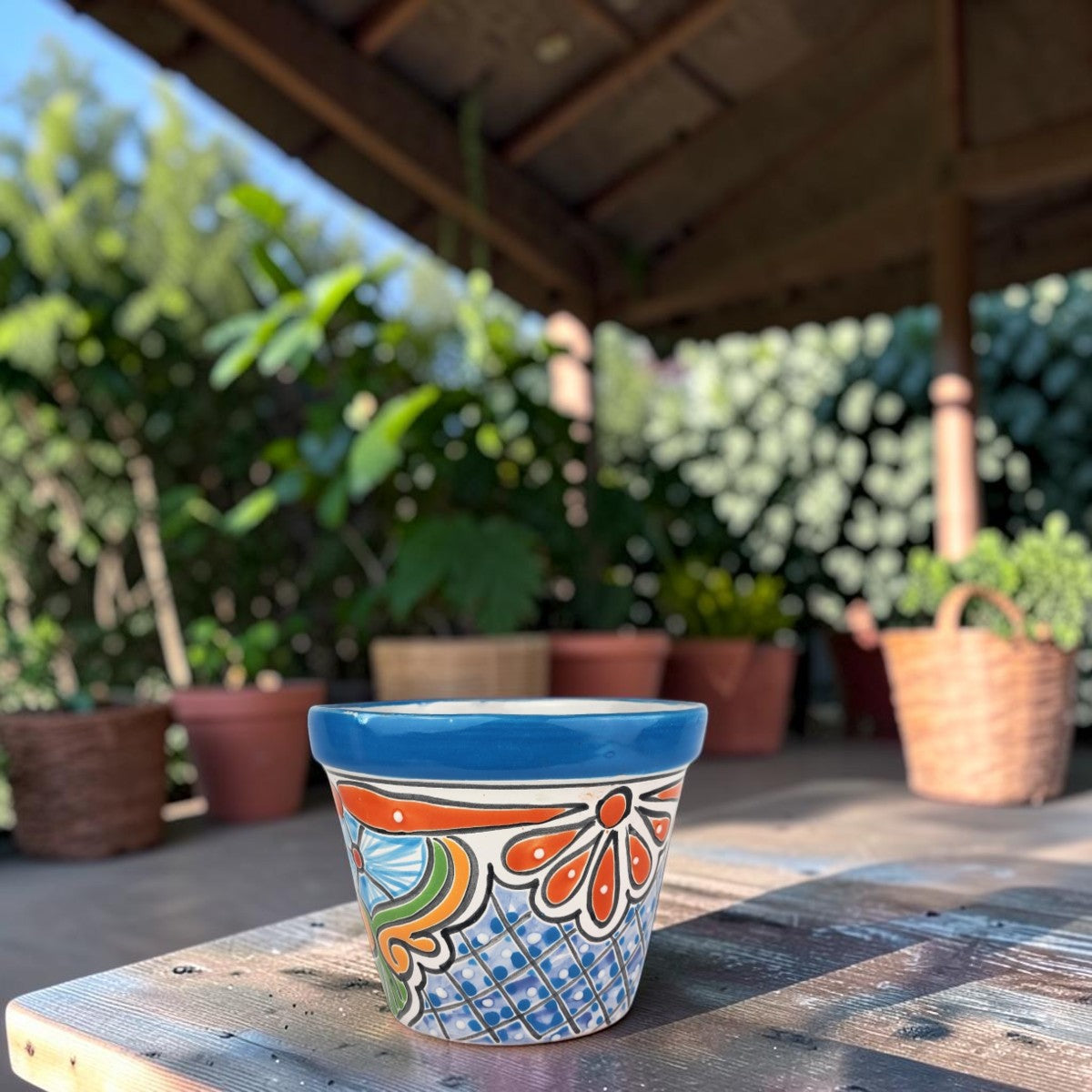 A small hand-painted ceramic planter pot with a blue rim and orange, green, and blue floral patterns, sitting on a wooden table in a garden patio setting.