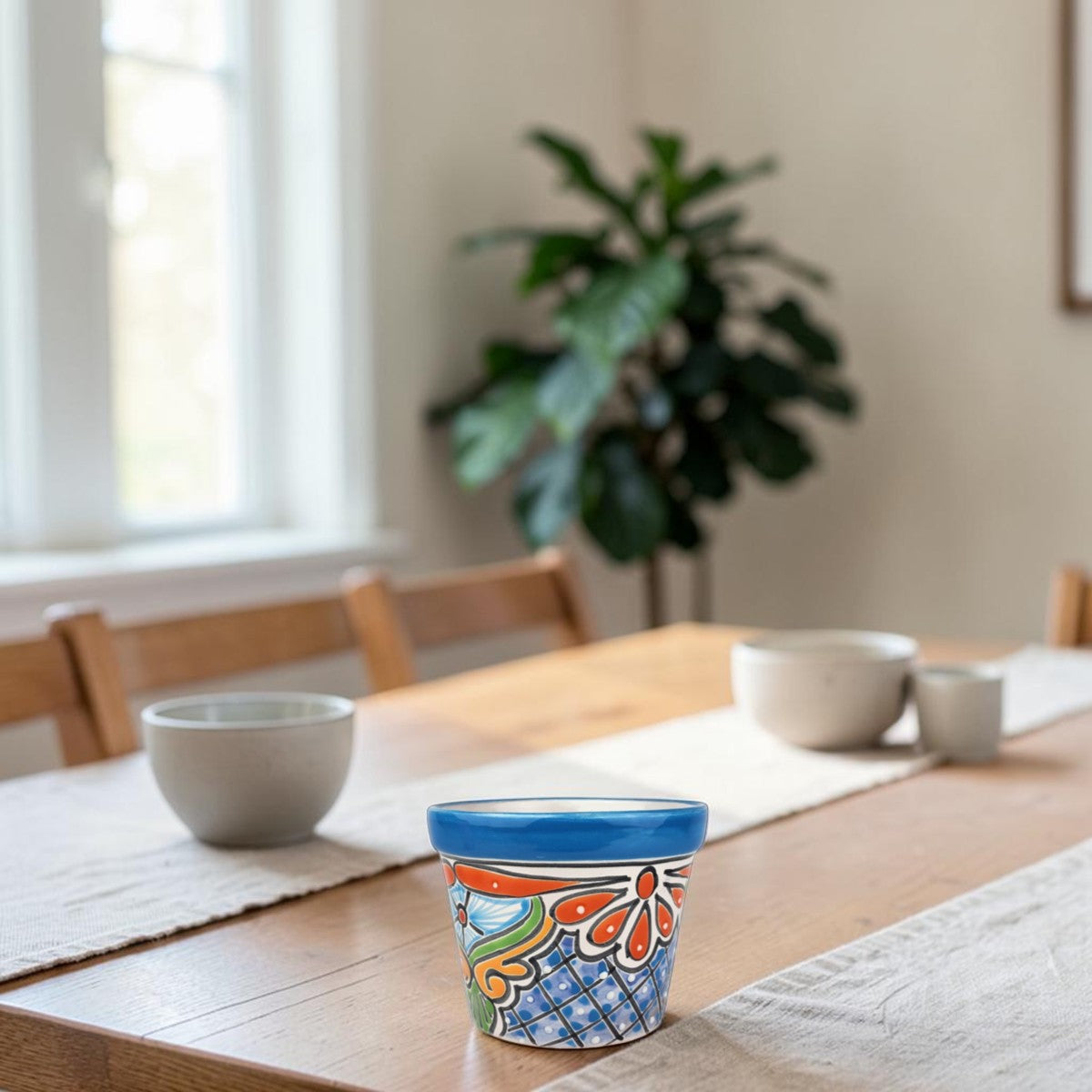A colorful ceramic planter pot with a blue rim and orange, green, and blue floral pattern sits on a wooden dining table in a bright, modern room with other ceramic bowls and a potted plant in the background.