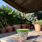 A small hand-painted ceramic planter pot with a green rim and colorful floral patterns, sitting on a wooden table in an outdoor patio setting with potted plants in the background.