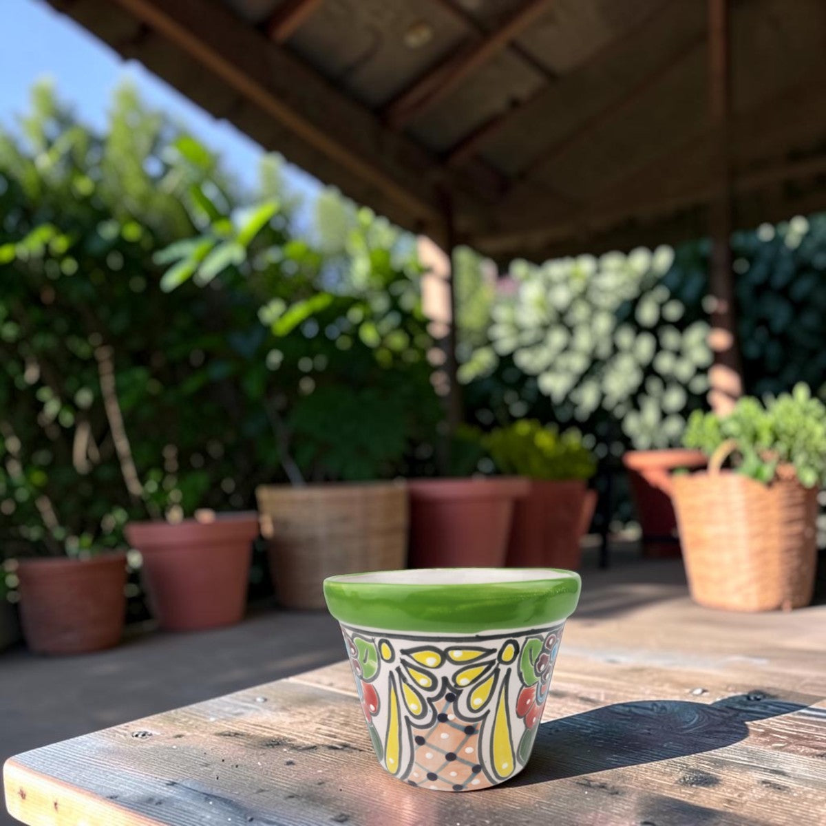 A small hand-painted ceramic planter pot with a green rim and colorful floral patterns, sitting on a wooden table in an outdoor patio setting with potted plants in the background.