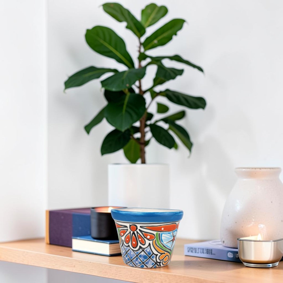 A colorful ceramic pot with a blue rim and orange, green, and blue floral pattern sits on a light wood shelf next to books, a white vase, and a candle, with a potted fiddle-leaf fig plant in the background.