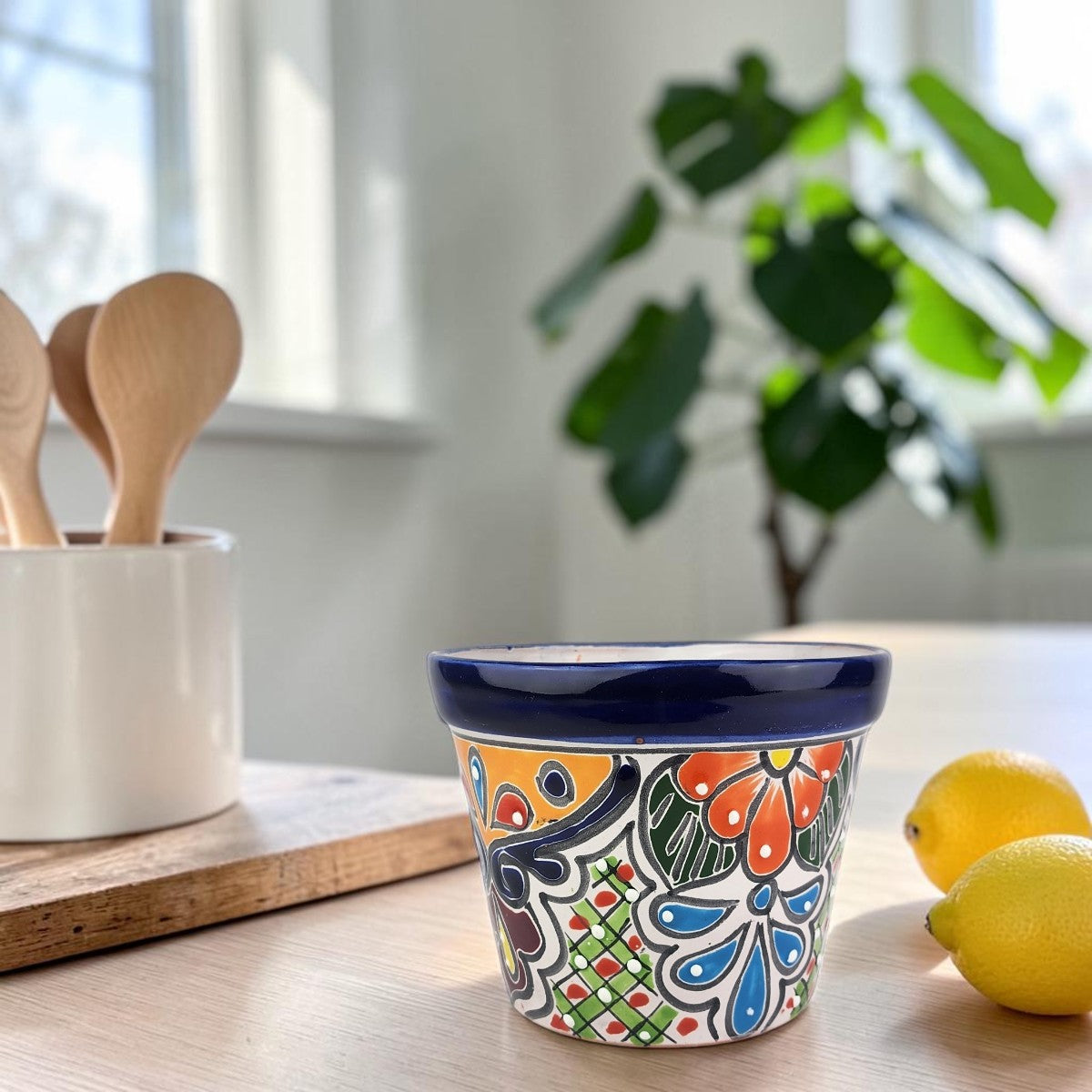 A small ceramic container with a colorful hand-painted floral pattern and a dark blue rim sits on a light wood table in a bright kitchen setting, with lemons and wooden spoons blurred in the background.