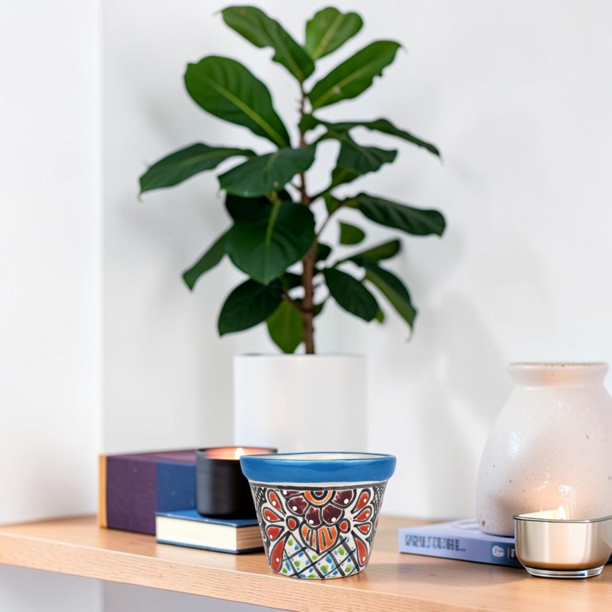 A small hand-painted ceramic pot with a blue rim and a colorful geometric floral pattern sits on a light wood shelf next to books, a white vase, and a candle, with a potted fiddle-leaf fig plant in the background.