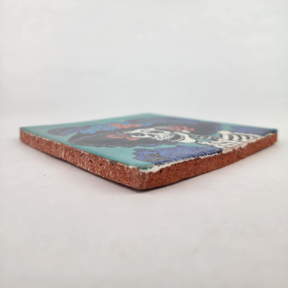 Angled view of teal Catrina Portrait ceramic tile showing glossy surface and natural terracotta edges