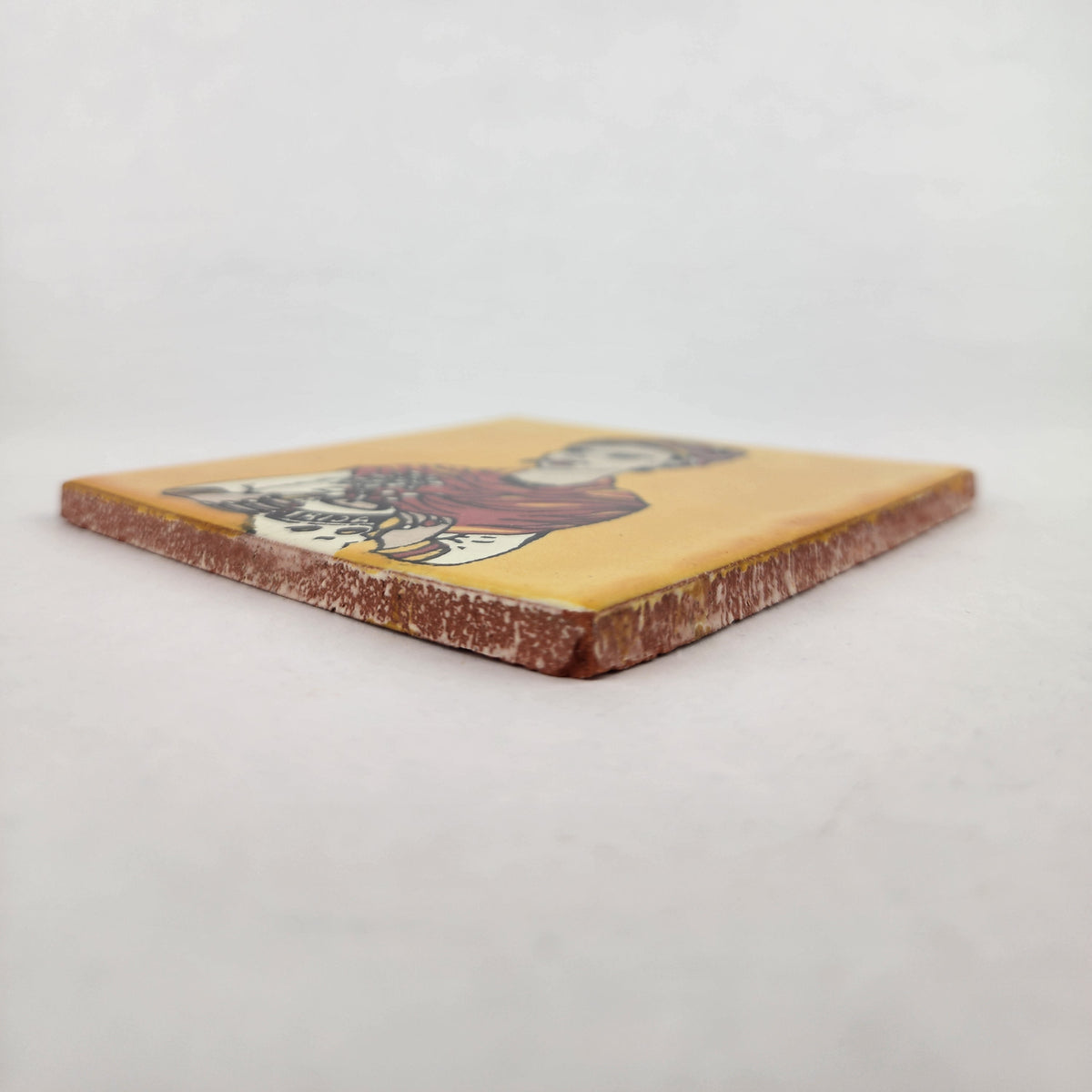 Angled view of mustard yellow Frida Kahlo portrait ceramic tile showing glossy surface and terracotta edges