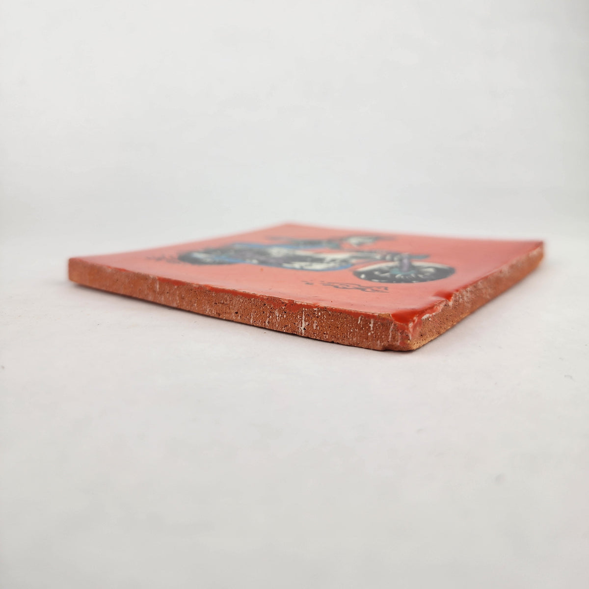 Angled view of Skeleton Motorcycle ceramic tile with red-orange background showing glazed surface and ceramic edge