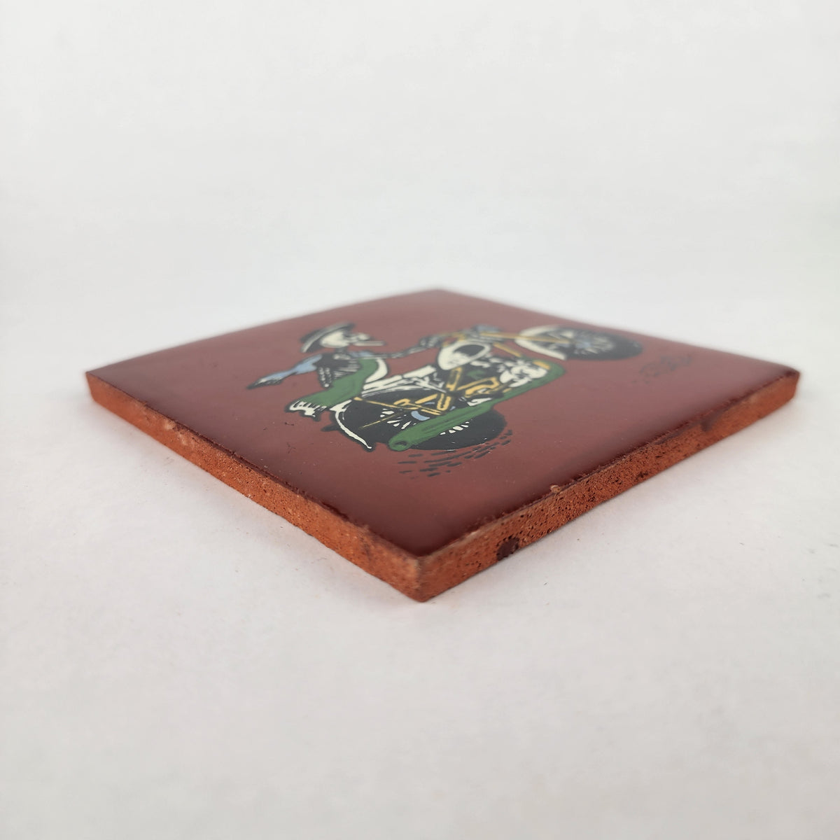 Angled view of Skeleton Motorcycle ceramic tile with red brown background showing glazed surface and ceramic edge