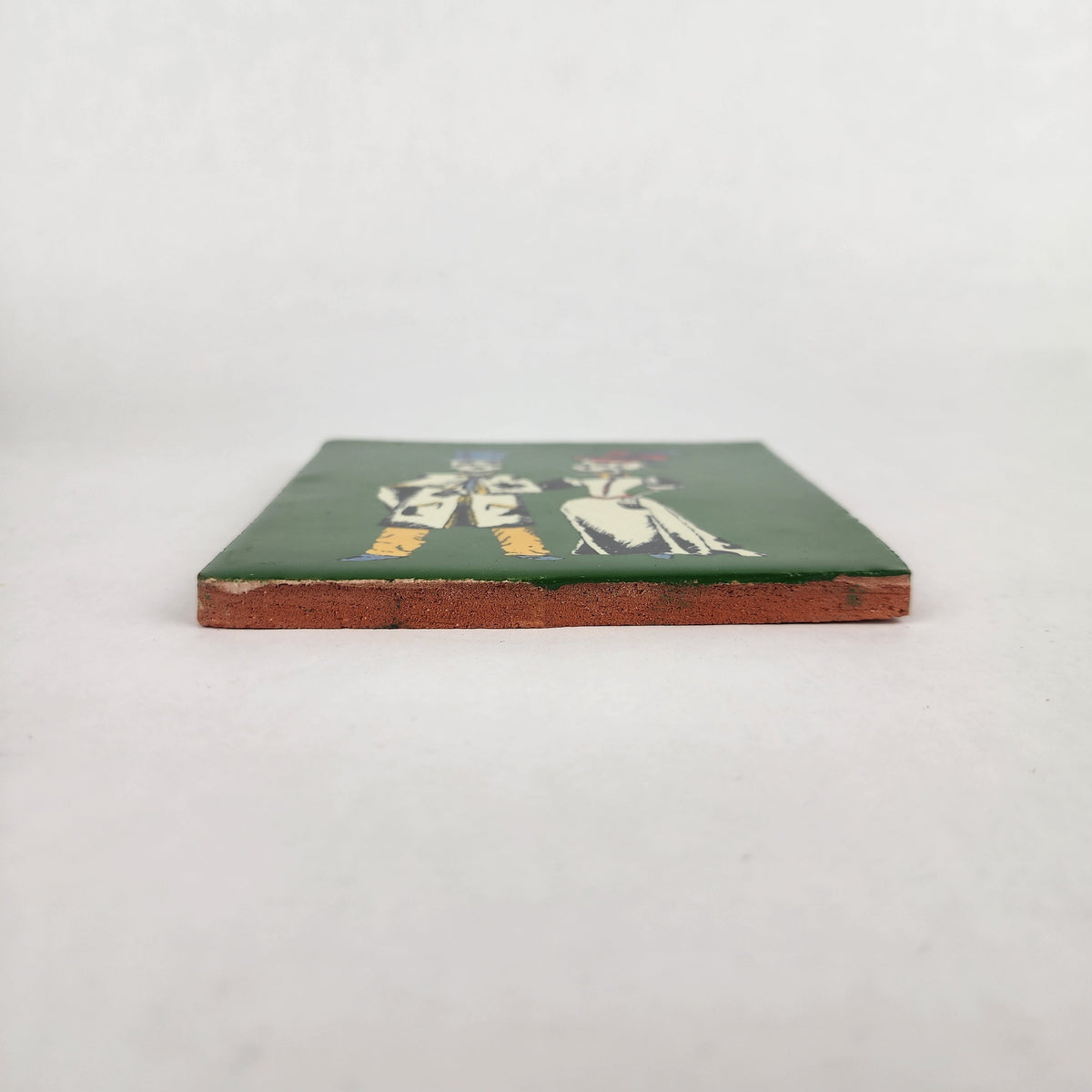 A small square ceramic tile with a dark green top, featuring a white illustration of two figures and a reddish-brown unglazed edge, photographed on a light gray background.
