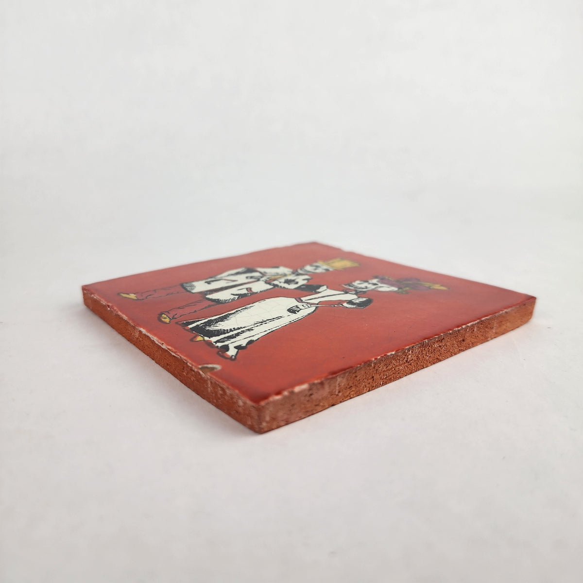 A square ceramic tile with a reddish-brown glaze, featuring an illustration of two figures in traditional clothing, set against a plain light gray background.