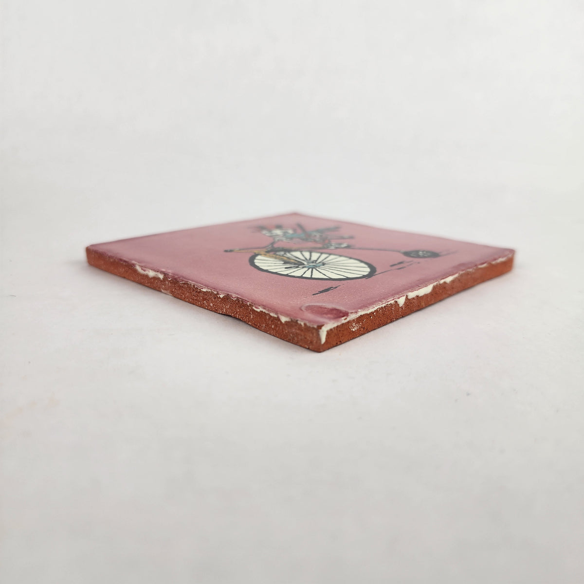 Angled view of pink skeleton bicycle ceramic tile showing glazed surface and ceramic edge
