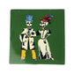 A square green ceramic tile featuring a vintage-style illustration of two skeleton figures dressed in formal attire, one in a blue top hat and coat with yellow trousers, and the other in a white dress with a red and blue feathered hat.