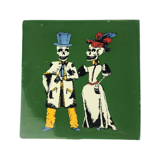 A square green ceramic tile featuring a vintage-style illustration of two skeleton figures dressed in formal attire, one in a blue top hat and coat with yellow trousers, and the other in a white dress with a red and blue feathered hat.