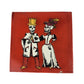 A square ceramic tile featuring a Day of the Dead (Dia de los Muertos) illustration of a skeleton bride and groom. The couple is dressed in traditional white and black attire, with the groom wearing a gold top hat and the bride a white gown with a purple and gold feathered hat.
