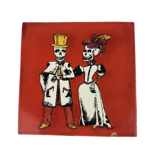 A square ceramic tile featuring a Day of the Dead (Dia de los Muertos) illustration of a skeleton bride and groom. The couple is dressed in traditional white and black attire, with the groom wearing a gold top hat and the bride a white gown with a purple and gold feathered hat.