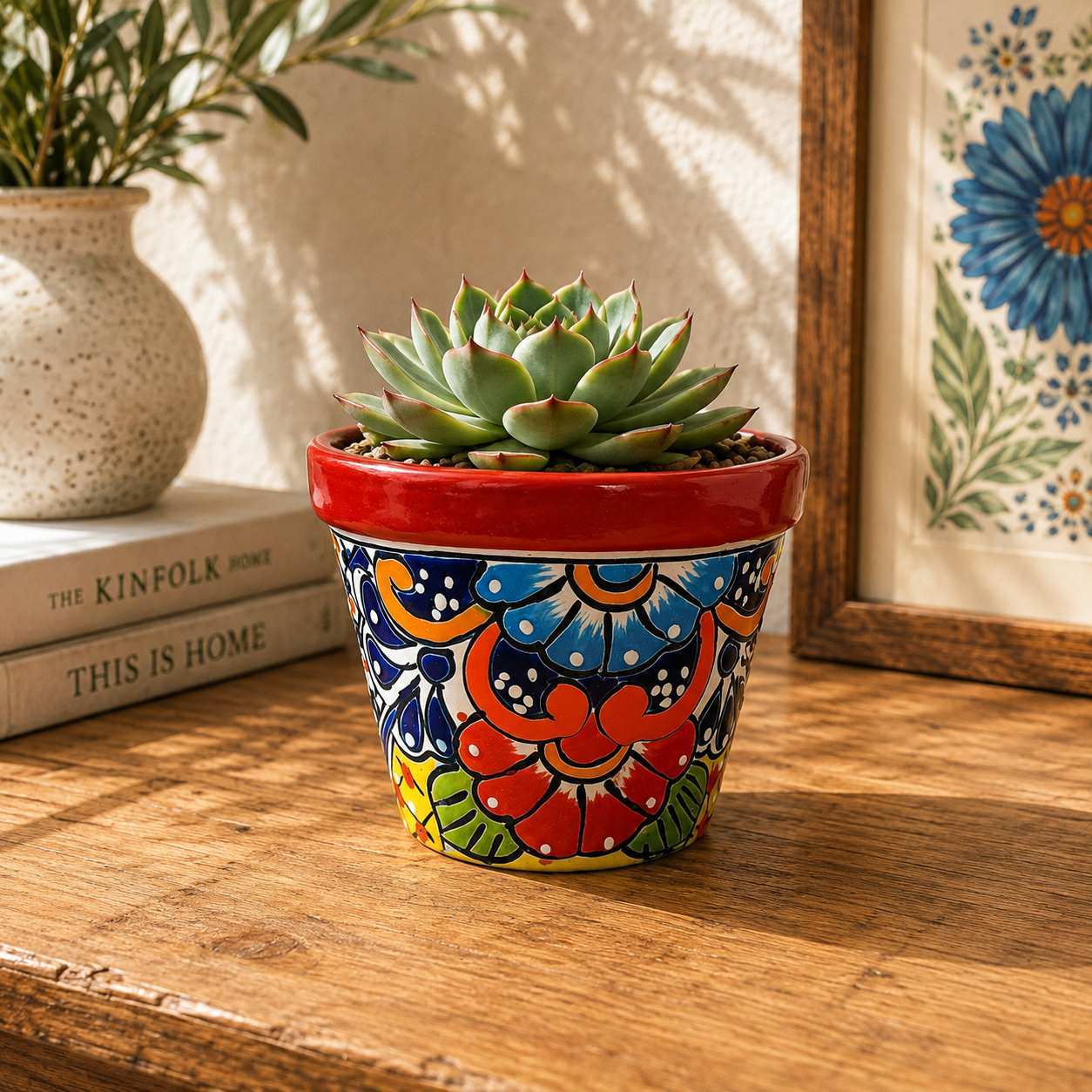 Red rim floral ceramic planter holding a succulent on a wooden table beside stacked books, a speckled vase, and framed blue botanical artwork.
