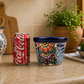 A decorated ceramic planter with a colorful floral pattern and a dark blue rim sits on a kitchen counter next to a red Coca-Cola can. The background features a tiled wall, a wooden cutting board, a potted green plant, and a bowl of limes.