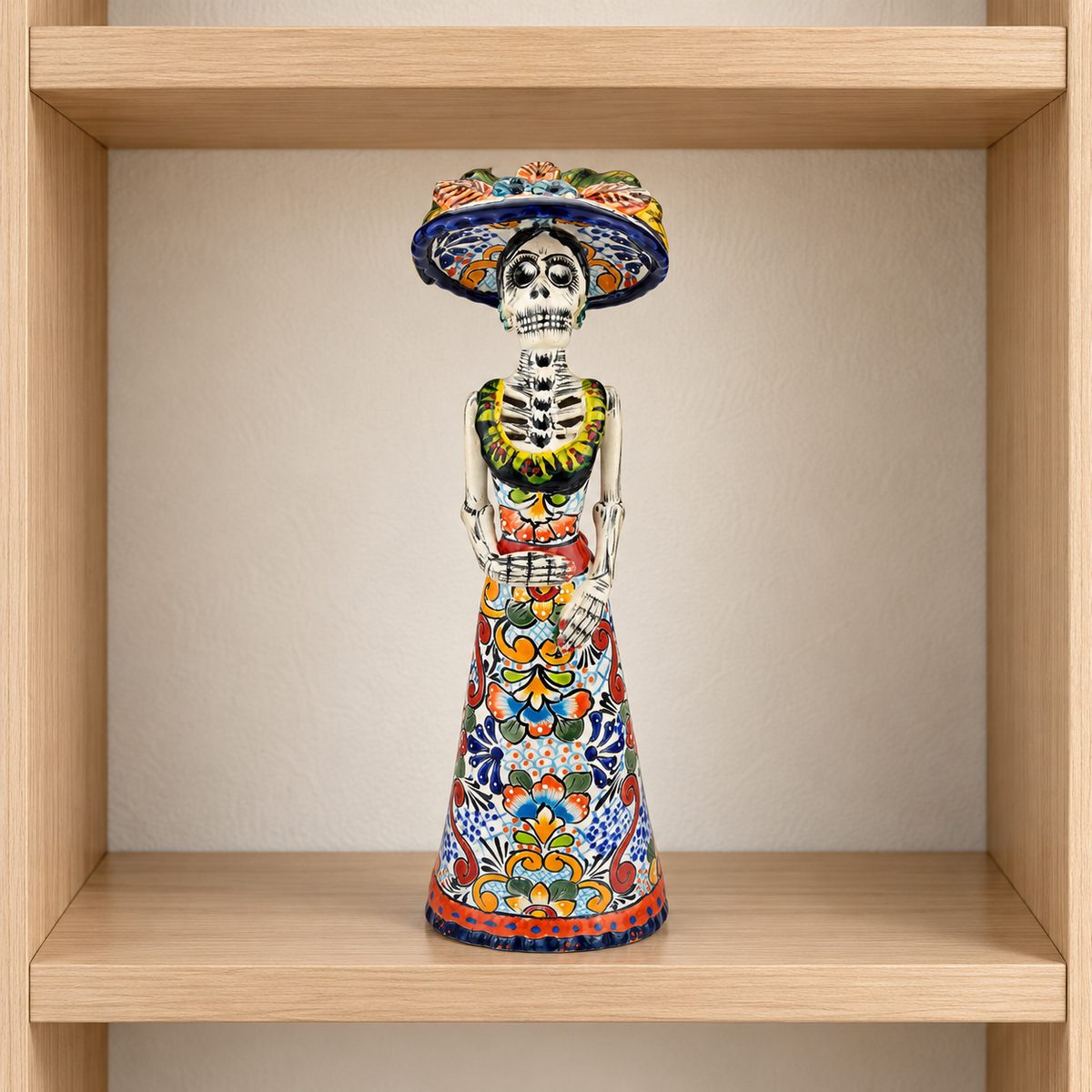 Colorful hand-painted ceramic Catrina figurine with a floral dress and blue sombrero displayed on a light wood shelf.