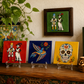 Three colorful Day of the Dead ceramic tiles on clear acrylic stands, displayed on a wooden mantel next to a potted plant and books, with a framed picture hanging on the wall above.