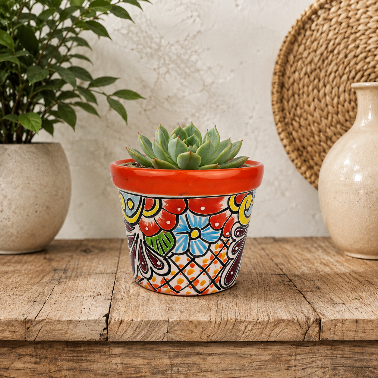 Red orange rim floral ceramic planter holding a succulent on a rustic wooden table beside neutral pottery and a woven wall decoration.