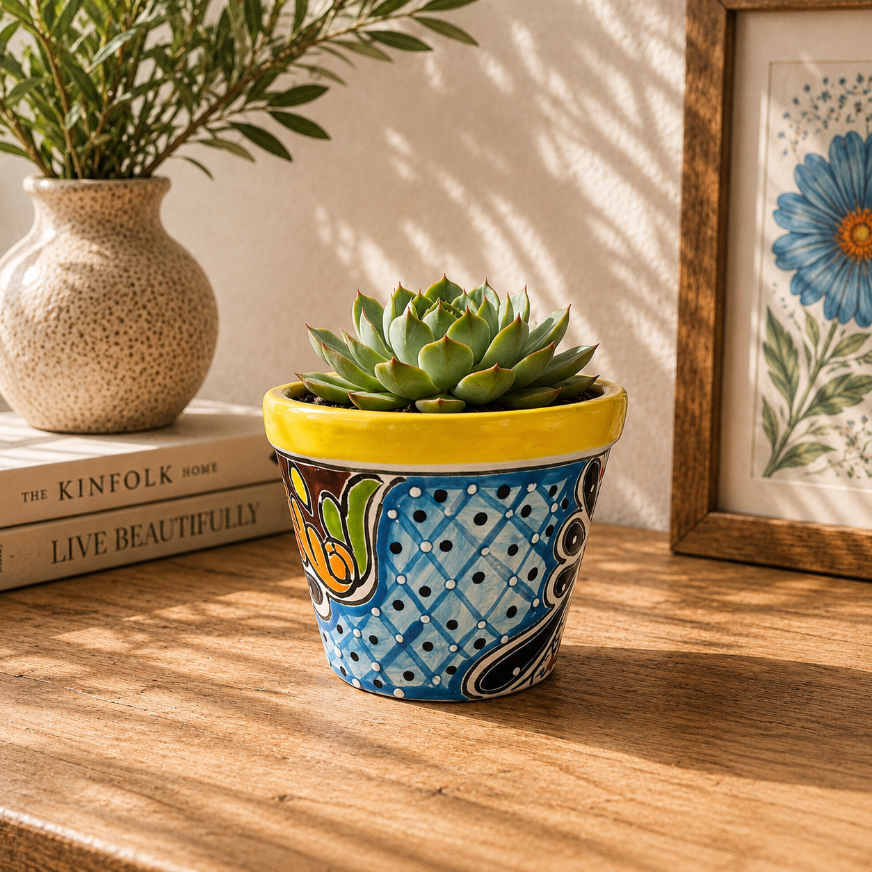 Yellow rim ceramic planter with blue lattice artwork holding a succulent on a wooden table beside books, a pottery vase, and botanical artwork.
