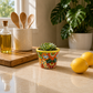 Yellow rim ceramic planter with orange flowers and green artwork holding a succulent on a sunlit kitchen counter beside lemons.