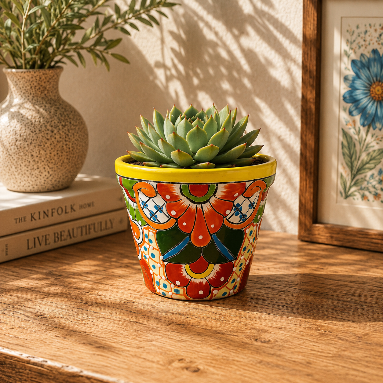 Yellow rim floral ceramic planter holding a succulent on a wooden table beside stacked books, a speckled vase, and framed botanical artwork.
