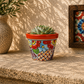 Red orange rim floral ceramic planter holding a succulent on a stone surface beside a speckled vase and framed floral tile artwork.
