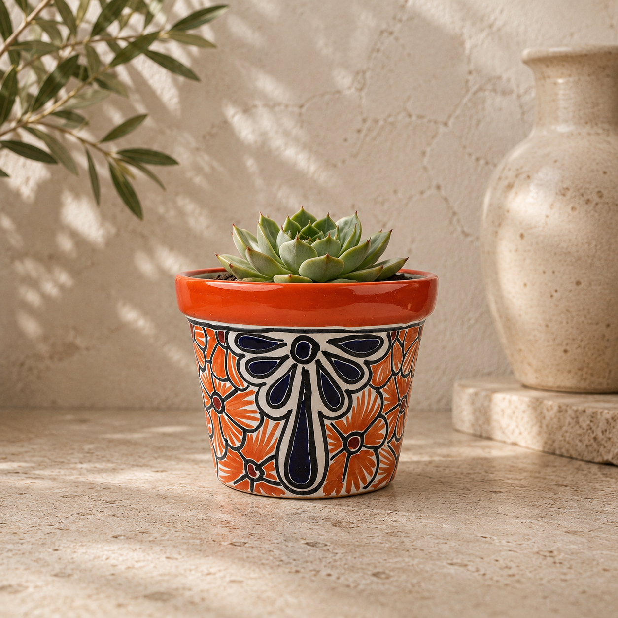 Red orange rim ceramic planter with orange and deep blue floral artwork holding a succulent on a stone surface beside a cream vase.
