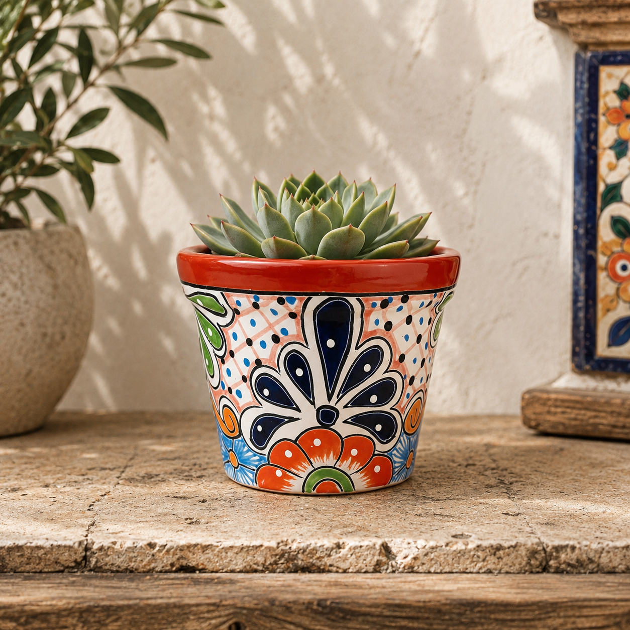 Red orange rim floral ceramic planter holding a succulent on a stone-topped table beside neutral pottery and framed decorative tile artwork.
