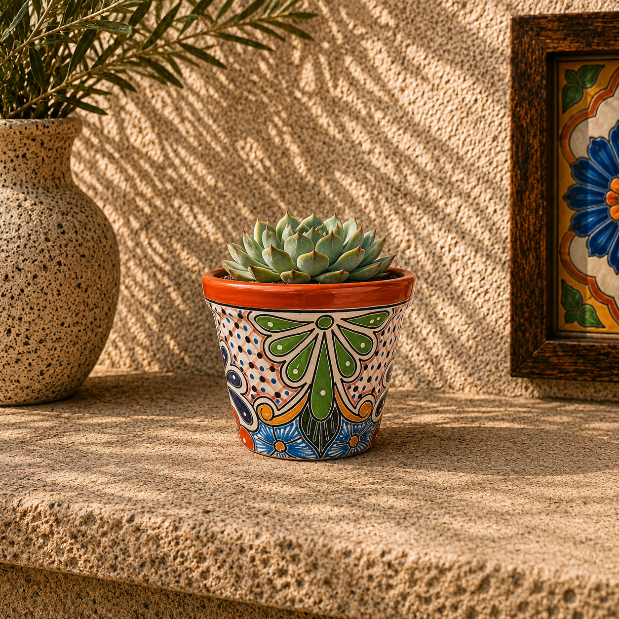 Red orange rim ceramic planter holding a succulent on a stone surface beside a speckled vase with leafy branches and framed blue floral tile artwork.
