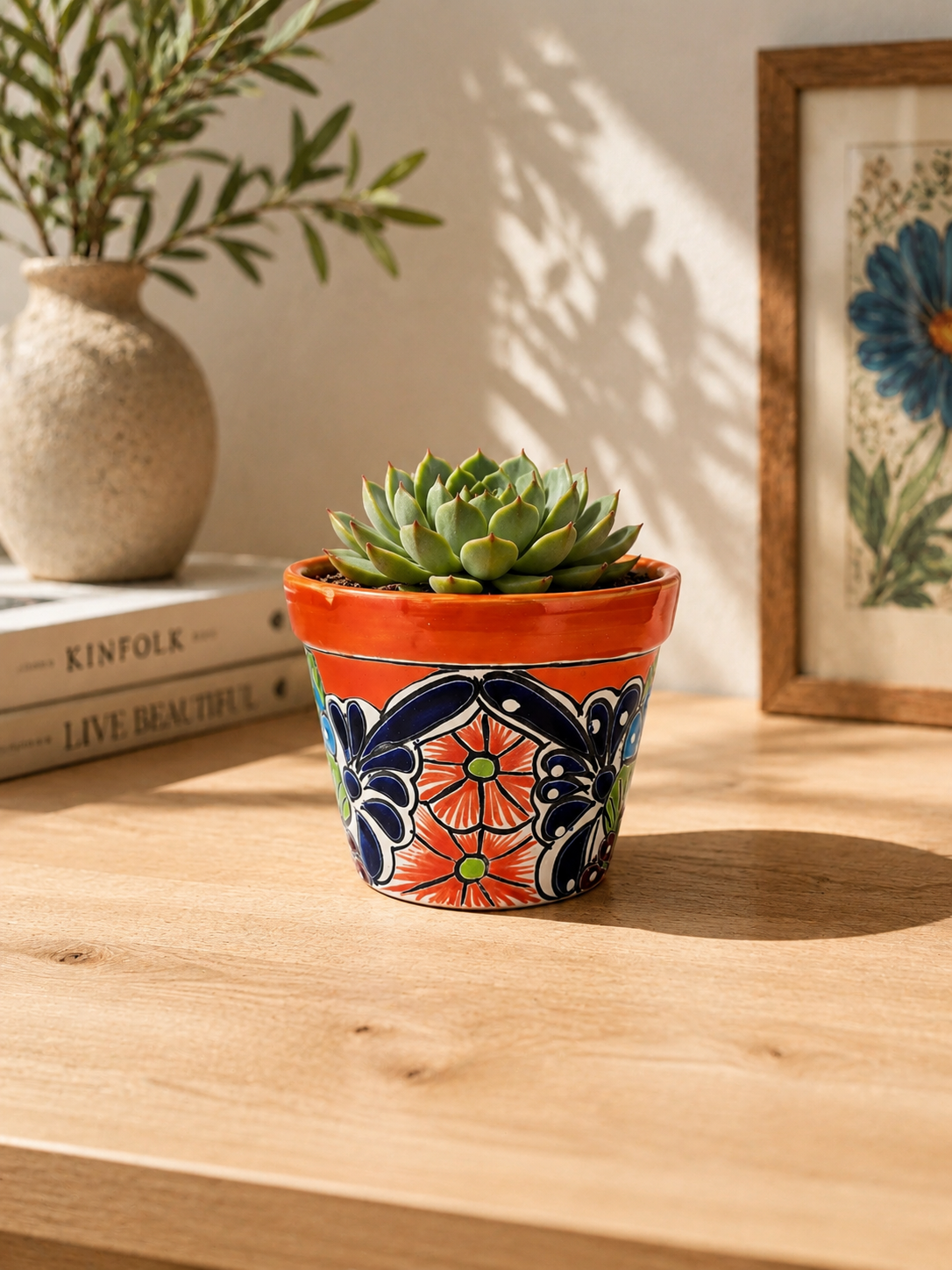 Red orange rim ceramic planter holding a succulent on a wooden table beside stacked books, a pottery vase, and framed botanical artwork.
