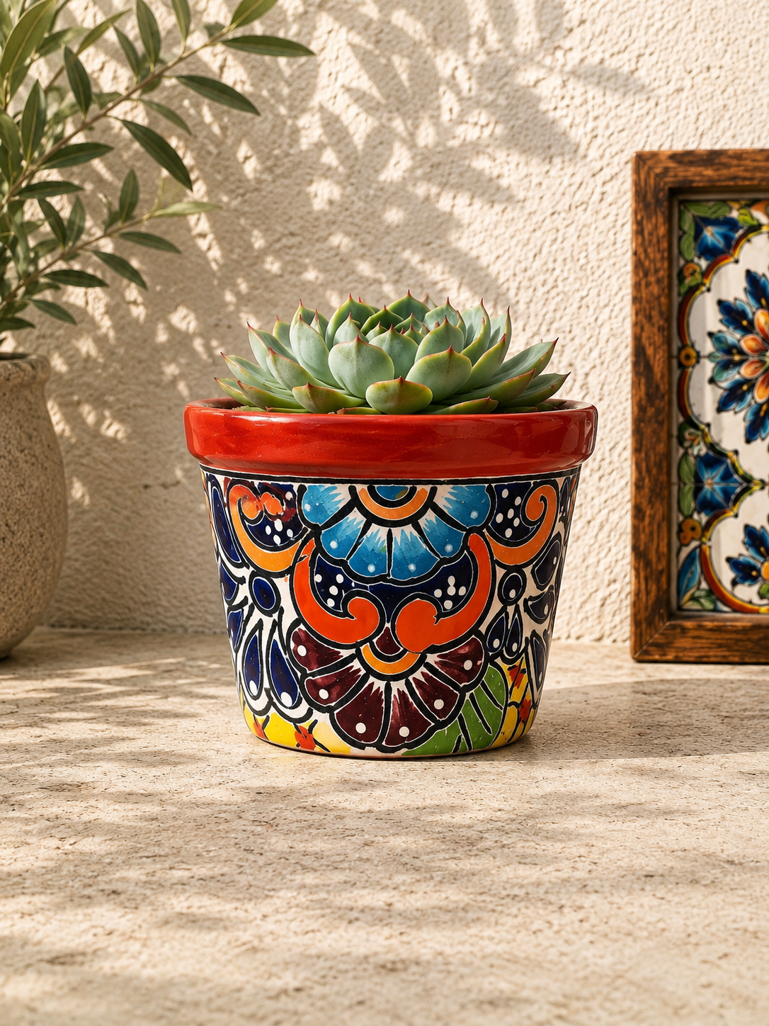 Red rim floral ceramic planter holding a succulent on a textured stone surface beside a potted tree and framed decorative tile artwork.
