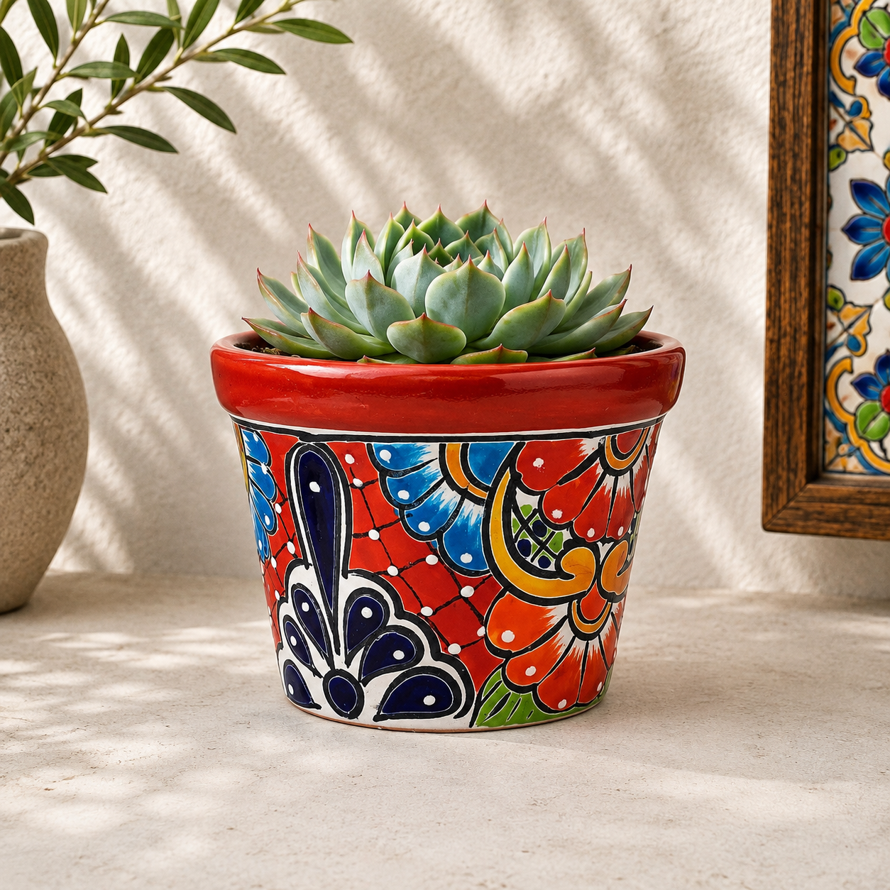 Red rim floral ceramic planter holding a succulent on a light stone surface beside neutral pottery and framed decorative tiles.
