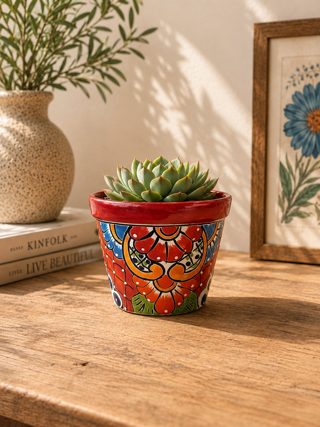 Red rim floral ceramic planter holding a succulent on a wooden table beside stacked books, a speckled vase, and framed botanical artwork.
