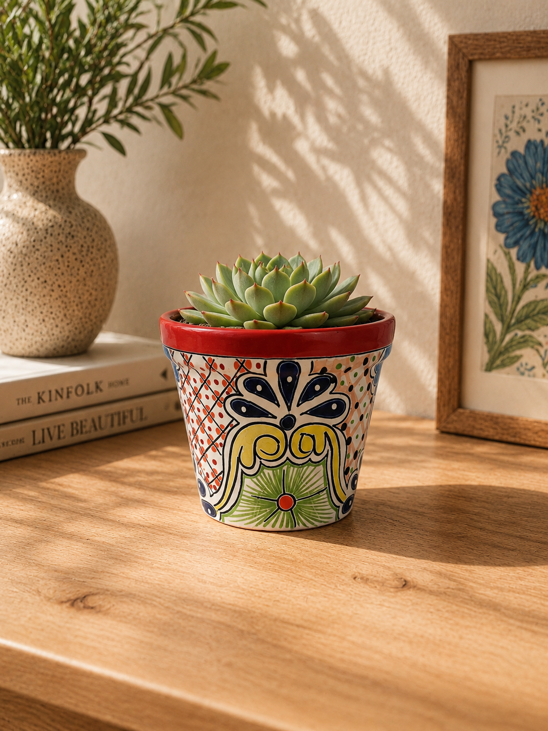Red rim ceramic planter holding a succulent on a wooden table beside stacked books, a speckled vase with leafy branches, and botanical artwork.
