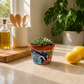 Red rim ceramic planter with sky blue flowers, deep blue petals, orange accents, and green leaf details on a white background.