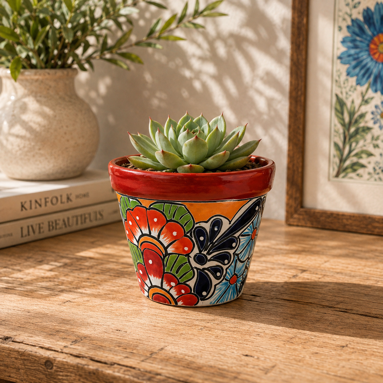 Red rim floral ceramic planter holding a succulent on a wooden table beside stacked books, a pottery vase with leafy branches, and botanical artwork.
