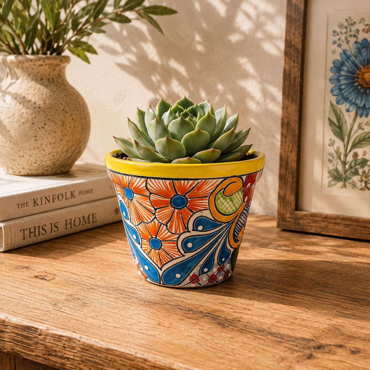 Yellow rim ceramic planter with orange and blue floral artwork holding a succulent on a wooden table beside books, pottery, and botanical artwork.
